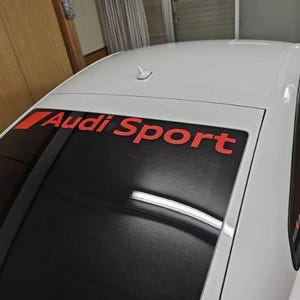 Op de afbeelding: Witte auto met een zwarte achterruit met de rode tekst "Audi Sport". Het dak van de auto heeft een kleine antenne. De afbeelding is genomen vanuit een iets verhoogde hoek.