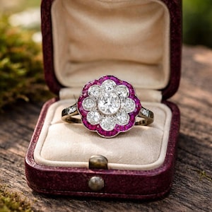 May include: A gold ring with a flower design, featuring a central diamond and surrounding rubies and smaller diamonds. The ring is presented in a burgundy box with a cream lining, ideal for gifting.