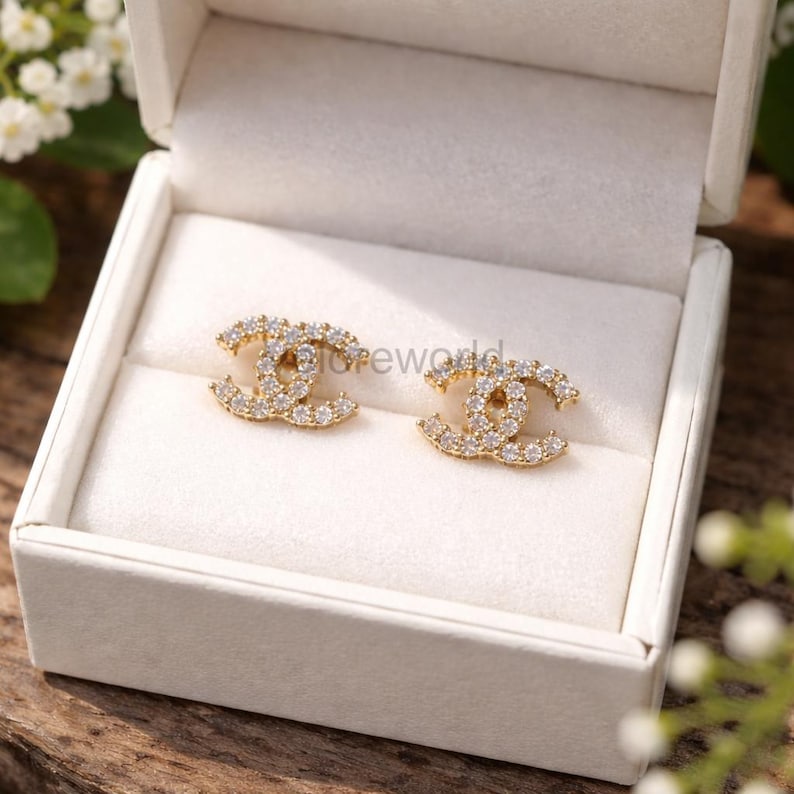 May include: A pair of gold-colored stud earrings in a double-C design, encrusted with small, clear crystals. The earrings are displayed inside a white jewelry box with a soft, white interior.
