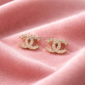 May include: A pair of gold-colored stud earrings in a double-C design, encrusted with small, clear crystals. The earrings are displayed on a pink, velvety surface, creating a luxurious aesthetic.