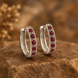 May include: A pair of silver hoop earrings featuring a row of small, round, deep red rubies set along the front face of each hoop. The earrings have a polished metallic finish and a hinged closure mechanism for secure wear.