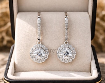 May include: A pair of silver drop earrings with a halo of small, sparkling stones surrounding a larger, central stone. The earrings are displayed in a black jewelry box with a cream-colored interior, set against a blurred background.