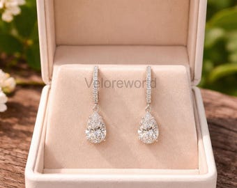 May include: A pair of elegant drop earrings with pear-shaped diamond-like stones, displayed in an open, cream-colored jewelry box. The earrings have a delicate, sparkling design, perfect for special occasions. The box is set against a natural background.