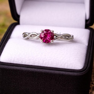 Può includere: Un anello d'argento con una vibrante pietra preziosa rosa, incastonato in un design a fascia intrecciata. L'anello è esposto in un cofanetto nero con interno bianco, creando un contrasto sorprendente. Il design dell'anello è elegante.