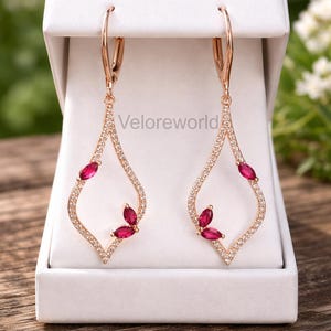 May include: Rose gold-tone earrings with a teardrop design. The earrings are adorned with small, sparkling crystals and three ruby-colored gemstones. The earrings are displayed in a white jewelry box.