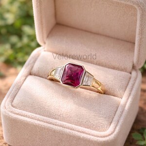 Peut inclure: Une bague en or avec une grande pierre précieuse rouge foncé rectangulaire au centre, flanquée de deux pierres précieuses claires rectangulaires plus petites. La bague est présentée dans un écrin beige. Le texte "Veloreworld" est visible.