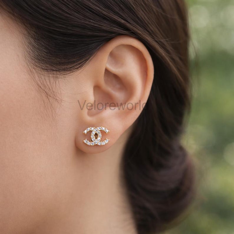 May include: A close-up of a gold-colored stud earring with a double-C design, encrusted with small, sparkling clear crystals. The earring is on a person's ear, with the brand name "Veloreworl" visible.