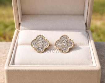 May include: A pair of gold-toned clover-shaped stud earrings, encrusted with small, sparkling crystals. The earrings are displayed inside an open, beige jewelry box, set against a soft, neutral interior. The box is on a surface with green foliage in the background.