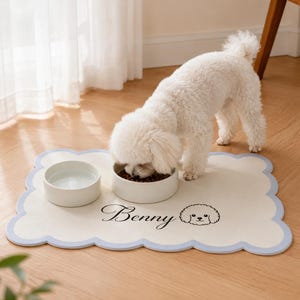 May include: A rectangular pet feeding mat with a scalloped light blue border and a cream-coloured surface. The mat features the name Benny in black script text next to a simple line-art illustration of a curly-haired dog's face.