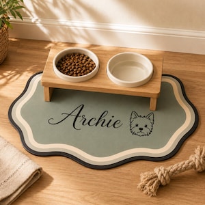 Puede incluir: Un comedero elevado de madera con dos cuencos de cerámica blanca, uno lleno de pienso marrón y otro con agua, sobre una alfombrilla para mascotas de color verde salvia con bordes ondulados, el nombre Archie en letra negra y un dibujo de un perro.