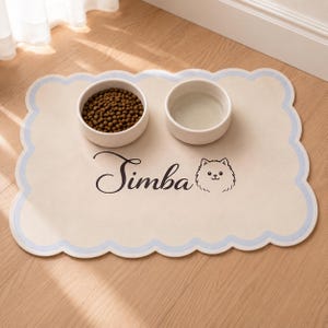 May include: A rectangular pet feeding mat with scalloped edges in light blue and cream. The mat features the name "Simba" in black cursive script next to a line-art illustration of a dog face. Two white ceramic bowls sit on top of the mat.