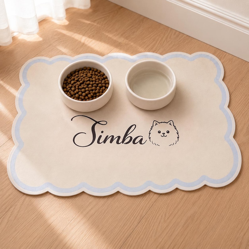 May include: A rectangular pet feeding mat with scalloped edges in light blue and cream. The mat features the name "Simba" in black cursive script next to a line-art illustration of a dog face. Two white ceramic bowls sit on top of the mat.