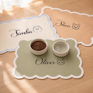 May include: Three rectangular pet feeding mats with scalloped edges in cream, tan, and sage green. Each mat features a custom name in black script and a small animal illustration. The sage green mat holds two white ceramic bowls for food and water.