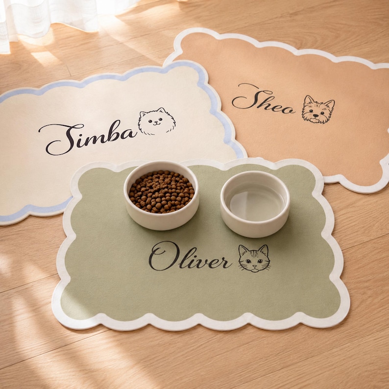 May include: Three rectangular pet feeding mats with scalloped edges in cream, tan, and sage green. Each mat features a custom name in black script and a small animal illustration. The sage green mat holds two white ceramic bowls for food and water.