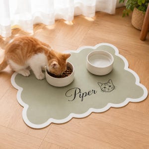 Puede incluir: Un tapete de alimentación para mascotas de color verde salvia con bordes festoneados blancos, que presenta el nombre Piper en letra cursiva negra y un dibujo lineal de una cara de gato. Dos cuencos de cerámica blanca están sobre el tapete.
