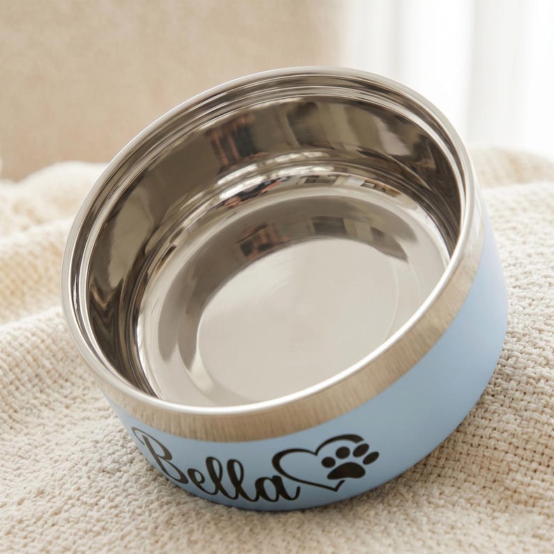 May include: A light blue stainless steel pet bowl with a polished silver interior. The exterior features the name Bella in black script lettering next to a black heart icon containing a paw print. The bowl has a cylindrical shape and a smooth finish.