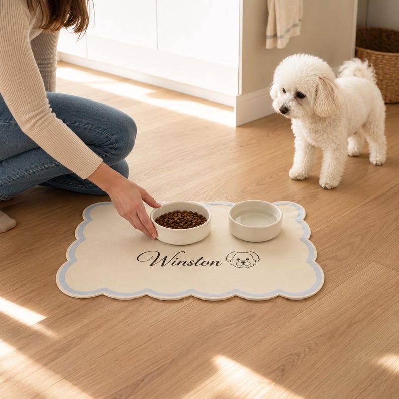 May include: A rectangular cream-colored pet feeding mat with a scalloped edge outlined in light blue. The mat features the name Winston in black script text alongside a simple line-art illustration of a dog face, holding two white bowls.