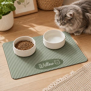 Pode incluir: Um tapete de alimentação retangular para animais de estimação com um padrão de riscas verticais verdes e brancas. O tapete apresenta um emblema oval verde com o nome Willow e um ícone de cara de gato impresso em branco. Duas taças de cerâmica.