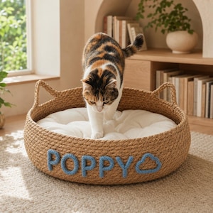 Puede incluir: Una cama redonda y tejida para mascotas de color tostado con lados en forma de orejas de gato y un cojín blanco suave. El exterior presenta el nombre "POPPY" y un icono de nube en letras de lana azul. Un gato calicó entra en la cama.