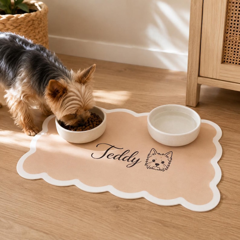 May include: A tan rectangular pet feeding mat with a scalloped white border, featuring the name Teddy and a line-art illustration of a dog. Two white ceramic bowls sit on the mat, one containing brown kibble and the other filled with water.