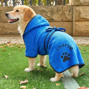 May include: A golden retriever wearing a blue dog robe with a hood and black trim. The robe has the name "Charlie" embroidered in script above a paw print. The dog is standing on green grass.