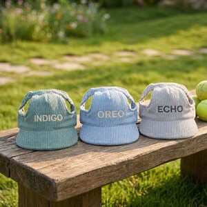 May include: Three corduroy hats in shades of green, blue, and gray, each with a different word embroidered on the front: "INDIGO", "OREO", and "ECHO". The hats are sitting on a wooden bench outdoors.