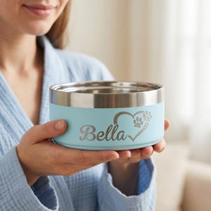 May include: A light blue pet bowl with a metallic silver rim. The side of the bowl features a laser-engraved design with the name Bella in a cursive script, accompanied by a heart shape that incorporates small paw prints into its outline.