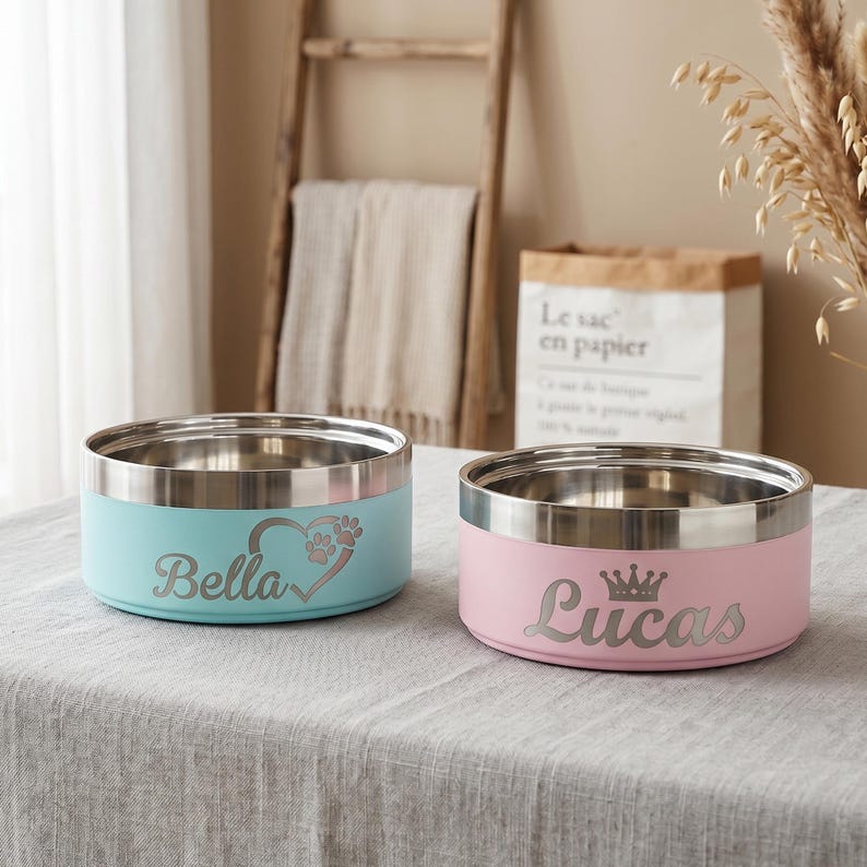 May include: Two round stainless steel pet bowls with a matte finish. The light blue bowl is engraved with the name Bella and a heart with a paw print. The light pink bowl is engraved with the name Lucas and a crown icon above the lettering.