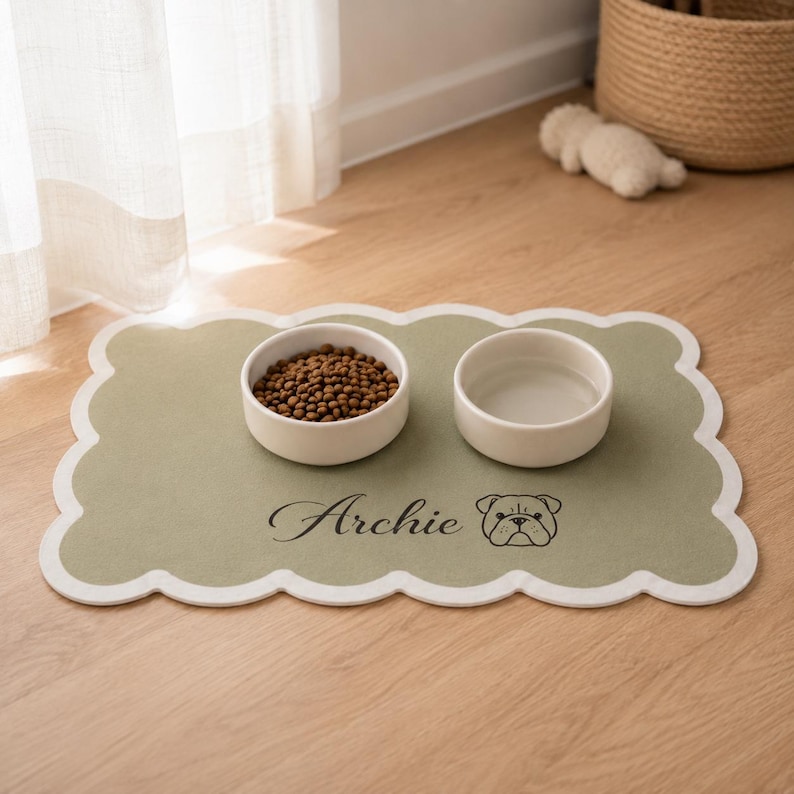 May include: A sage green rectangular pet feeding mat with a scalloped white border. The mat features the name Archie in black script text next to a black line drawing of a bulldog face. Two white ceramic bowls sit on top, one filled with brown kibble.