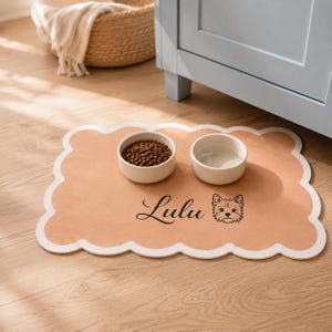 May include: A tan scalloped pet feeding mat with a white border, featuring the name Lulu in black script and a black line drawing of a dog face. Two round white ceramic bowls sit on the mat, one filled with brown dry pet food.