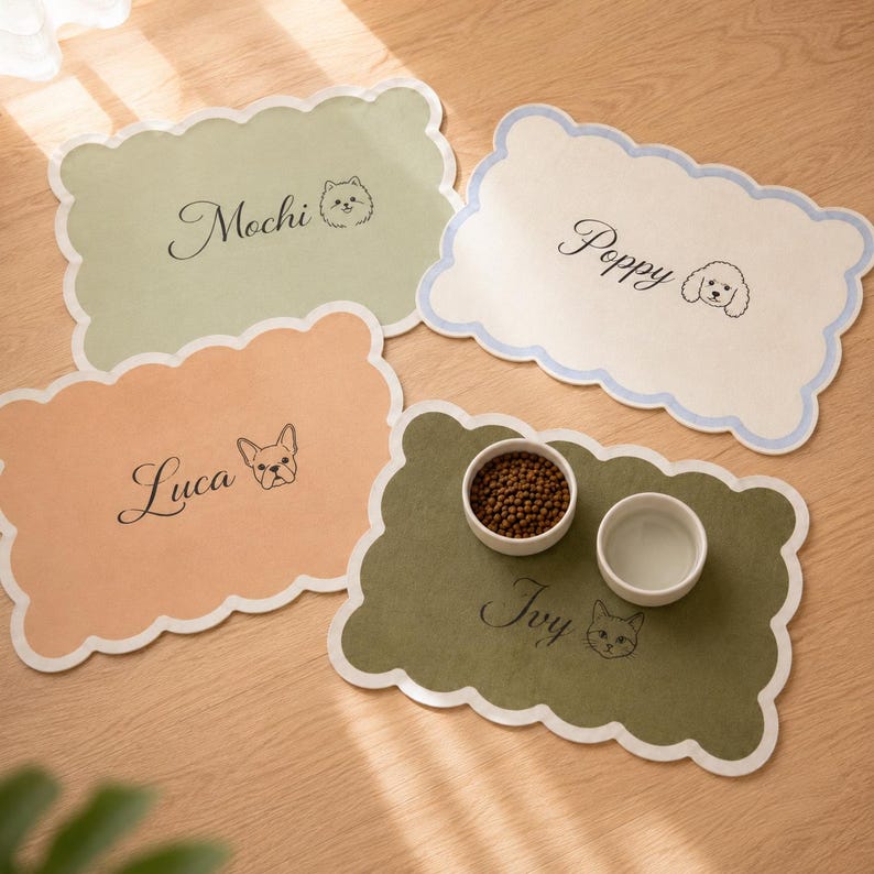 May include: Four rectangular pet placemats with scalloped edges in sage green, cream, peach, and olive green. Each mat features a custom name in cursive script and a line-art animal illustration. One olive mat holds two white bowls with food and water.