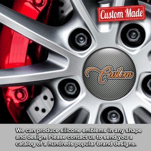 May include: Close-up of a silver car wheel with a red brake caliper. The wheel features a custom emblem with the word "Custom" in orange script. A red banner reads "Custom Made".