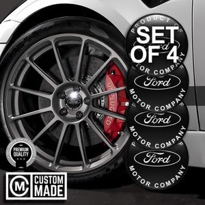 May include: A set of four black Ford Motor Company wheel centre caps. The caps have a white Ford logo and text. The image also shows a car wheel with a red brake caliper and the text "UN PLUG ED". The image also has the text "CUSTOM MADE".