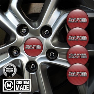 Może przedstawiać: Zbliżenie felgi samochodowej ze srebrnym wykończeniem. Koło posiada czerwoną osłonę piasty z napisem "YOUR WHEEL BRAND HERE" w kolorze srebrnym. Dodatkowe czerwone osłony są wyświetlane po prawej stronie. Logo "CUSTOM MADE" znajduje się w lewym dolnym rogu.