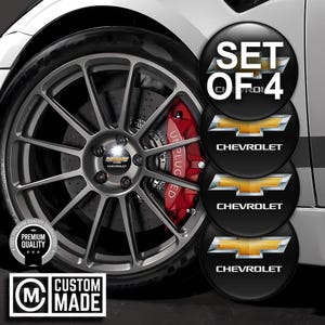 May include: A set of four black Chevrolet wheel center caps with the Chevrolet logo in gold. The caps are displayed next to a car wheel with a red brake caliper. The text "SET OF 4" is visible.