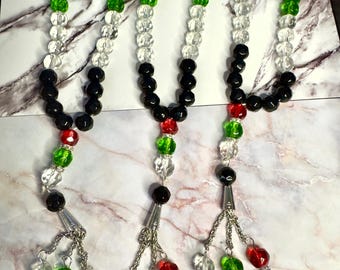 May include: Three beaded necklaces featuring red, green, black, and clear beads. Each necklace has a silver charm with dangling beads and a flag-shaped pendant. The beads are arranged in a colour pattern.