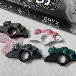May include: Four dragon-shaped rings in black, white, and green. Each ring features a detailed dragon figurine resting on a curved band. The rings are displayed on a soft, white surface. The book "Onyx Storm" is visible in the background.