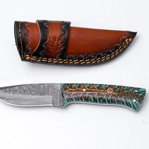 May include: A handcrafted knife with a patterned steel blade and a handle featuring teal, brown, and gold accents. The knife is paired with a brown leather sheath with decorative stitching.