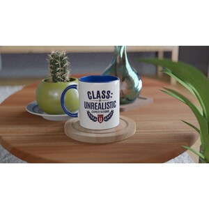 Op de afbeelding: Een witte keramische mok met een blauwe binnenkant en handvat. De voorzijde bevat donkerblauwe tekst met de tekst CLASS: OF UNREALISTIC EXPECTATIONS, met een klein rood en wit schildlogo onderaan gecentreerd tussen twee donkerblauwe lauriertakken.