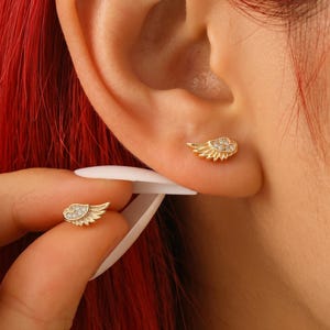 以下が含まれることがあります： 小さな透明な石が付いた、金色の翼型のスタッドピアス。片方のイヤリングは白いツールで持ち上げられ、もう片方は耳に装着されています。イヤリングは繊細で詳細なデザインです。