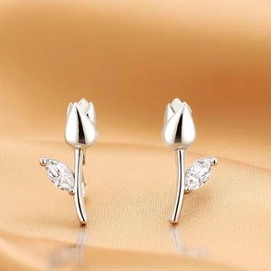 Op de afbeelding: Een paar zilverkleurige tulp oorknopjes. Elke oorbel heeft een tulpbloem met een helder, marquise-geslepen edelsteenblad. De oorbellen zijn geplaatst tegen een goudbruine, getextureerde achtergrond.