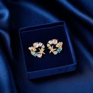 Könnte beinhalten: Ein Paar goldfarbene Ohrringe in einer blauen Samtbox. Jeder Ohrring hat ein kreisförmiges Design mit einem zentralen hellrosa Stein, umgeben von klaren und blauen Kristallen und kleinen Seestern-Akzenten.