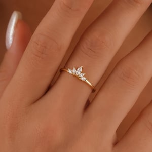 Può includere: Un delicato anello d'oro con un design a corona. L'anello è adornato con diverse pietre preziose chiare taglio marquise. L'anello è mostrato sul dito di una persona, evidenziando la sua estetica elegante e raffinata.
