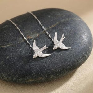 Puede incluir: Un par de pendientes plateados con forma de pájaro. Cada pendiente presenta un diseño detallado de golondrina, suspendido de una delicada cadena. Los pendientes se exhiben sobre una superficie de piedra oscura y texturizada, mostrando su intrincada artesanía.
