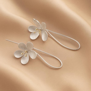 May include: A pair of silver-toned earrings featuring a floral design. Each earring has a flower with five petals and a small gold-colored center. The earrings have a long, curved wire that extends from the flower.