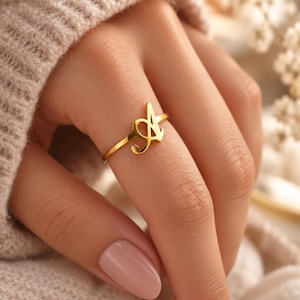 Puede incluir: Un anillo de tono dorado que presenta una letra A estilizada y prominente en una fuente cursiva. La banda es delgada y lisa, rodeando el dedo. El diseño se centra en la letra mayúscula A, que sirve como punto focal central de la pieza de joyería.