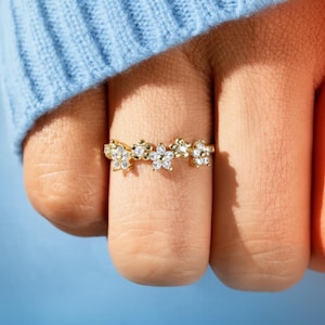 Puede incluir: Un anillo dorado con un diseño floral con pequeñas piedras preciosas transparentes. El anillo está compuesto por múltiples elementos en forma de flor, cada uno adornado con piedras brillantes. El anillo se muestra en un dedo, con un suéter azul en el fondo.