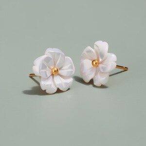 Könnte beinhalten: Ein Paar weiße Ohrstecker in Blütenform mit goldfarbenen Stiften und Zentren. Die Ohrringe haben einen perlmuttartigen Glanz. Das florale Design ist vor einem gedämpften grünen Hintergrund platziert. Ein zartes Accessoire.