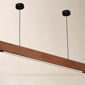 May include: A linear pendant light fixture featuring a long, rectangular dark brown wooden beam with a built-in LED light strip on the underside. The fixture is suspended from the ceiling by two thin black cables attached to two black circular mounts.