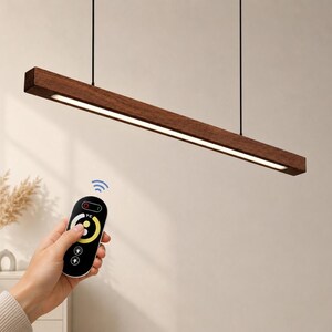 May include: A linear pendant light fixture made of dark brown wood with an integrated LED light strip. The fixture is suspended by two thin black cables. A black remote control with a circular yellow interface and power icons is shown.
